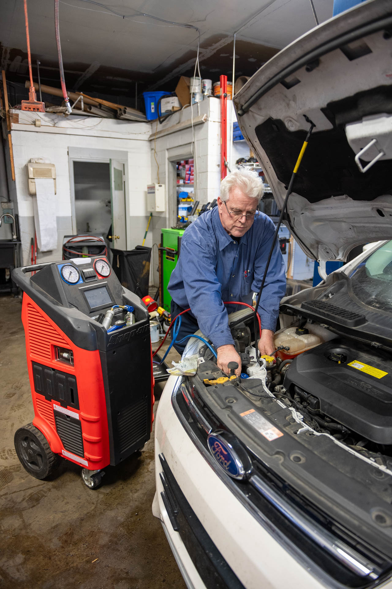 An older male mechanic repairs a car engine in a cluttered workshop, with tools and equipment surrounding him.