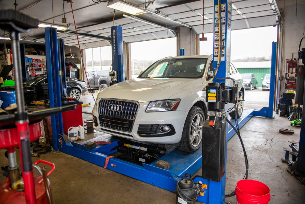 A white audi elevated on a lift in an auto repair garage, surrounded by various tools and equipment.