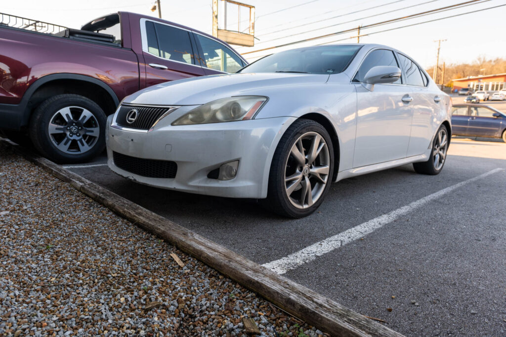 A silver lexus sedan parked in a gravel lot beside a maroon suv, with a wooden parking block in front.