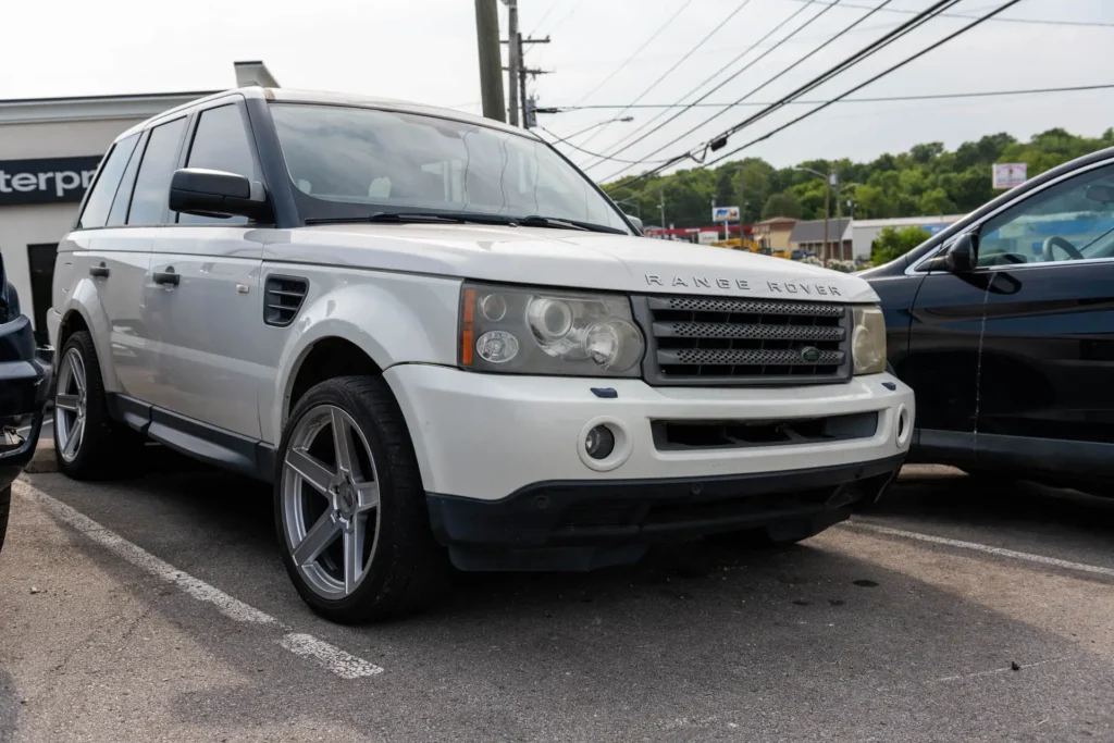 A white Range Rover is parked in a lot, surrounded by other vehicles. The car has tinted windows and silver alloy wheels. Utility poles and wires are visible in the background.