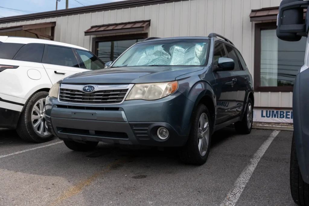 A parked blue Subaru Forester with a sunshade on the windshield is positioned in a parking lot next to a white vehicle.