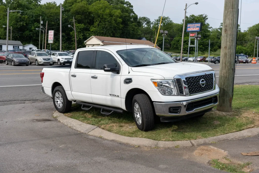 White pickup truck parked on a grass verge near a roadside, next to a utility pole, with a background of trees, other parked cars, and road signs.