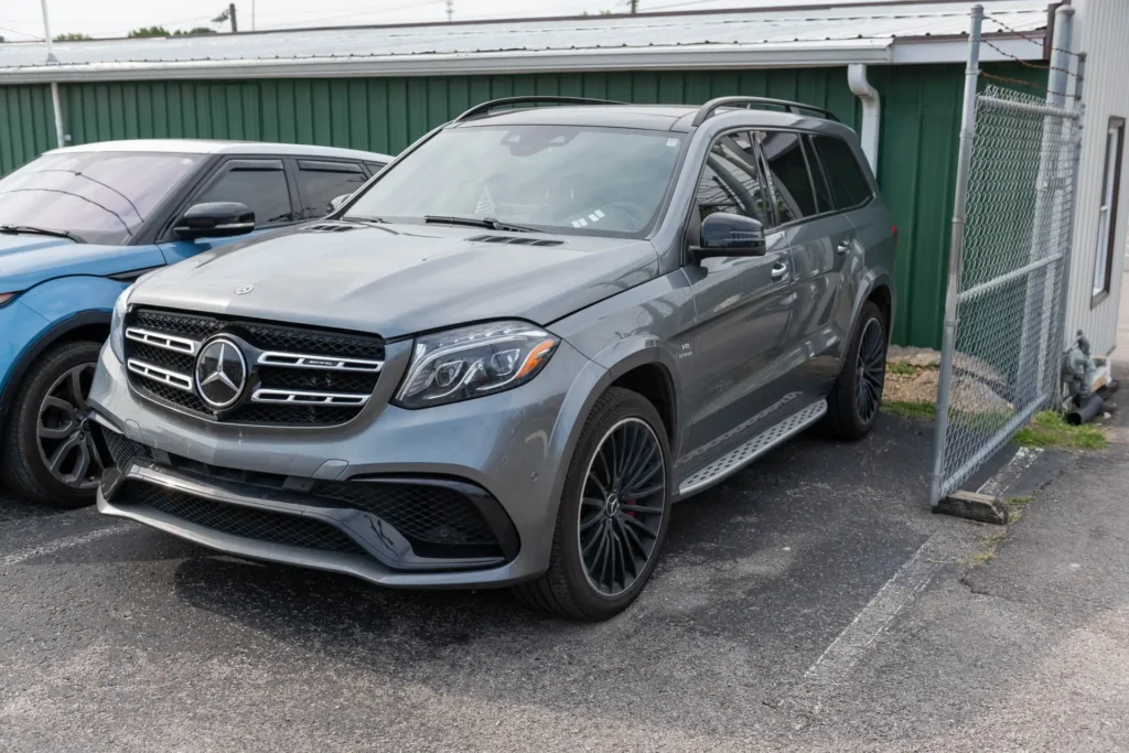 A silver Mercedes-Benz SUV is parked outside next to a blue car and a green building.