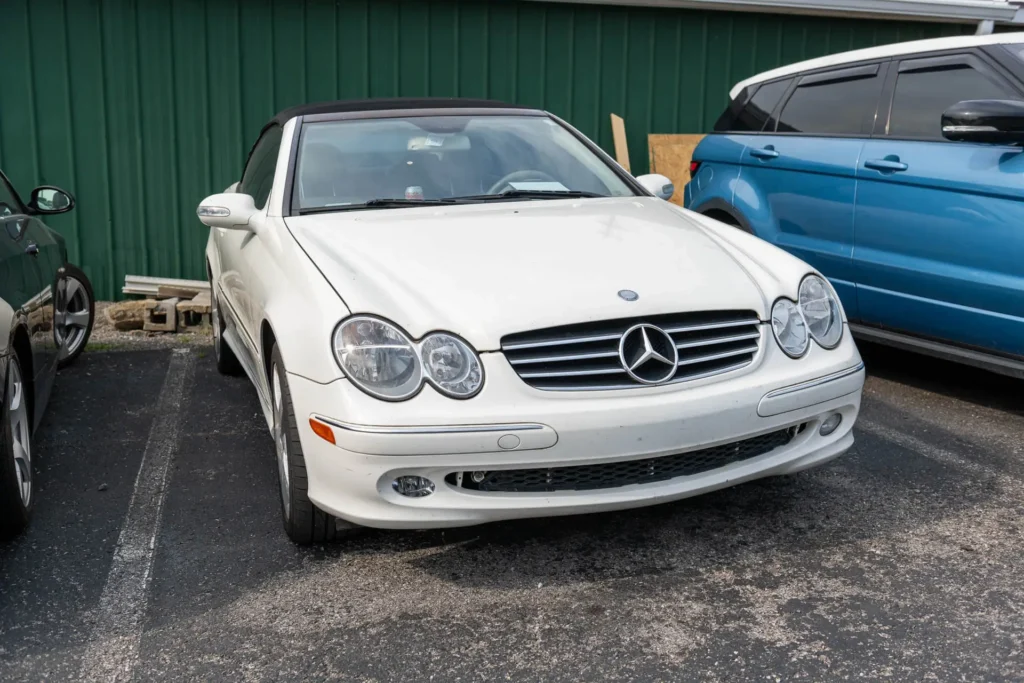 A white Mercedes-Benz convertible is parked in a lot next to a blue and a gray vehicle near a green wall.