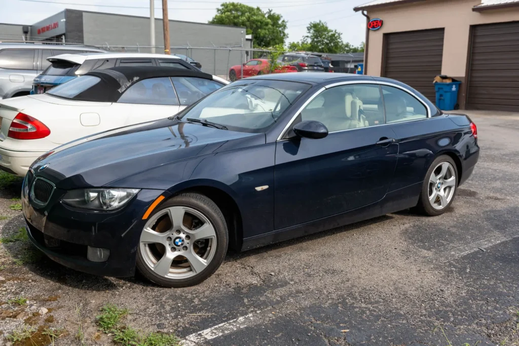 A parked blue BMW convertible is shown with the roof up, positioned in a parking lot alongside other vehicles.