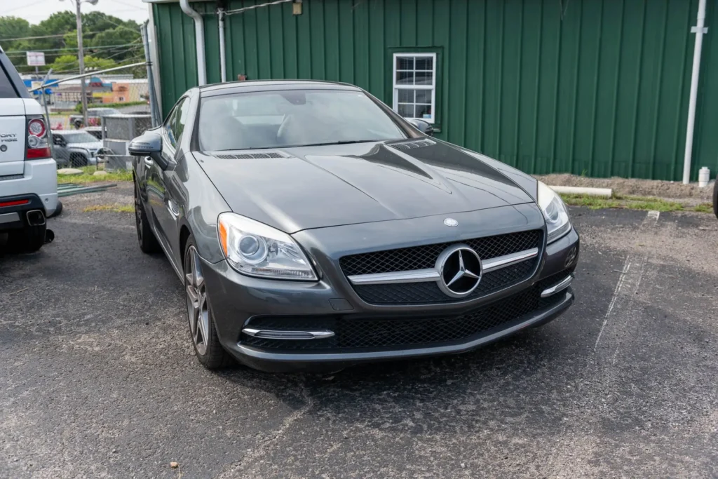 A dark gray Mercedes-Benz sports car is parked on an asphalt surface in front of a green building.