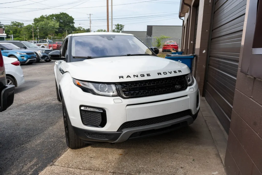 A white Range Rover SUV parked next to a brown garage door on a paved surface in an urban area, with other vehicles and buildings visible in the background.