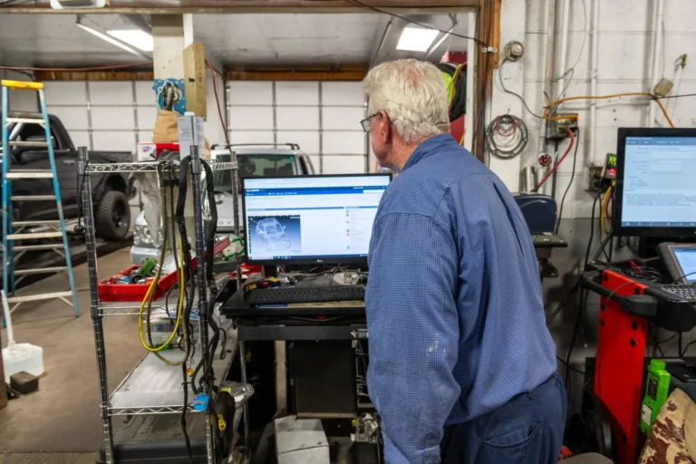 A person with gray hair in a blue shirt is standing in front of a computer screen in a workshop with various tools and equipment.